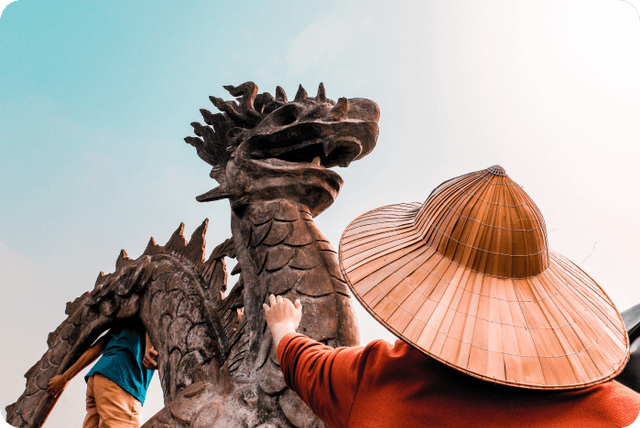 A person wearing a broad-brimmed hat reaches out with one hand to touch a rough, weathered-looking stone dragon.