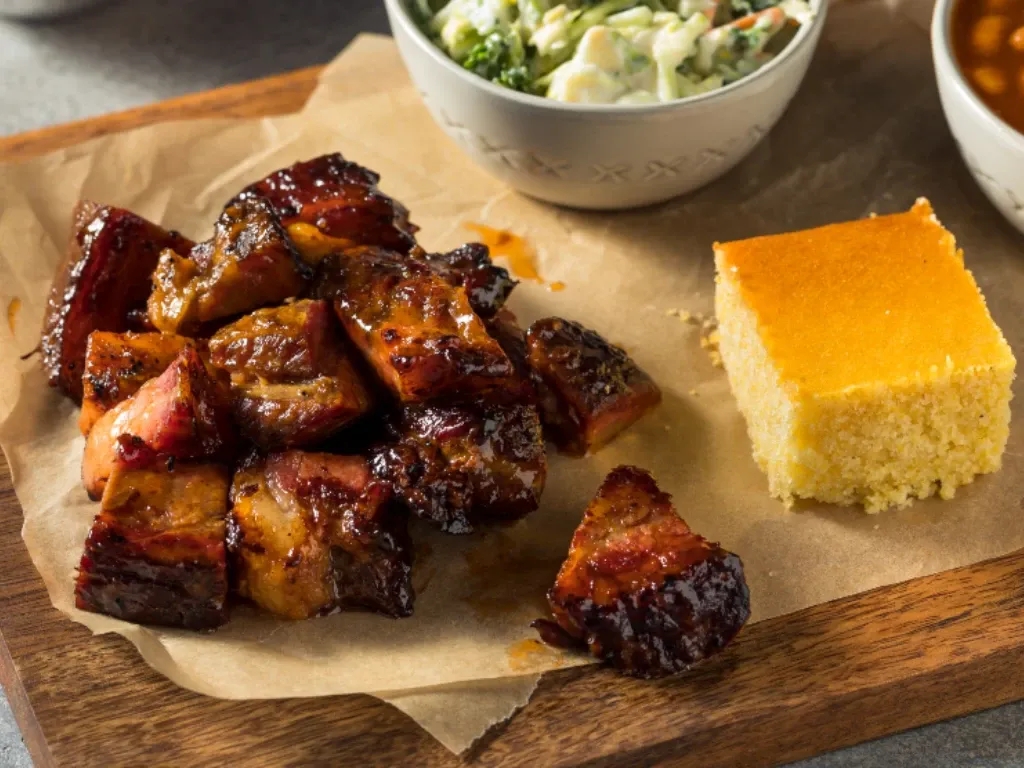 Saucy barbecued burnt ends sit on butcher paper with sides of cornbread and baked beans