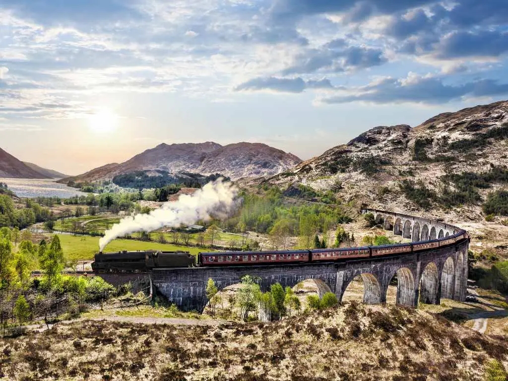 Glenfinnan Railway Viaduct in Scotland with the Jacobite steam train against sunset over lake.